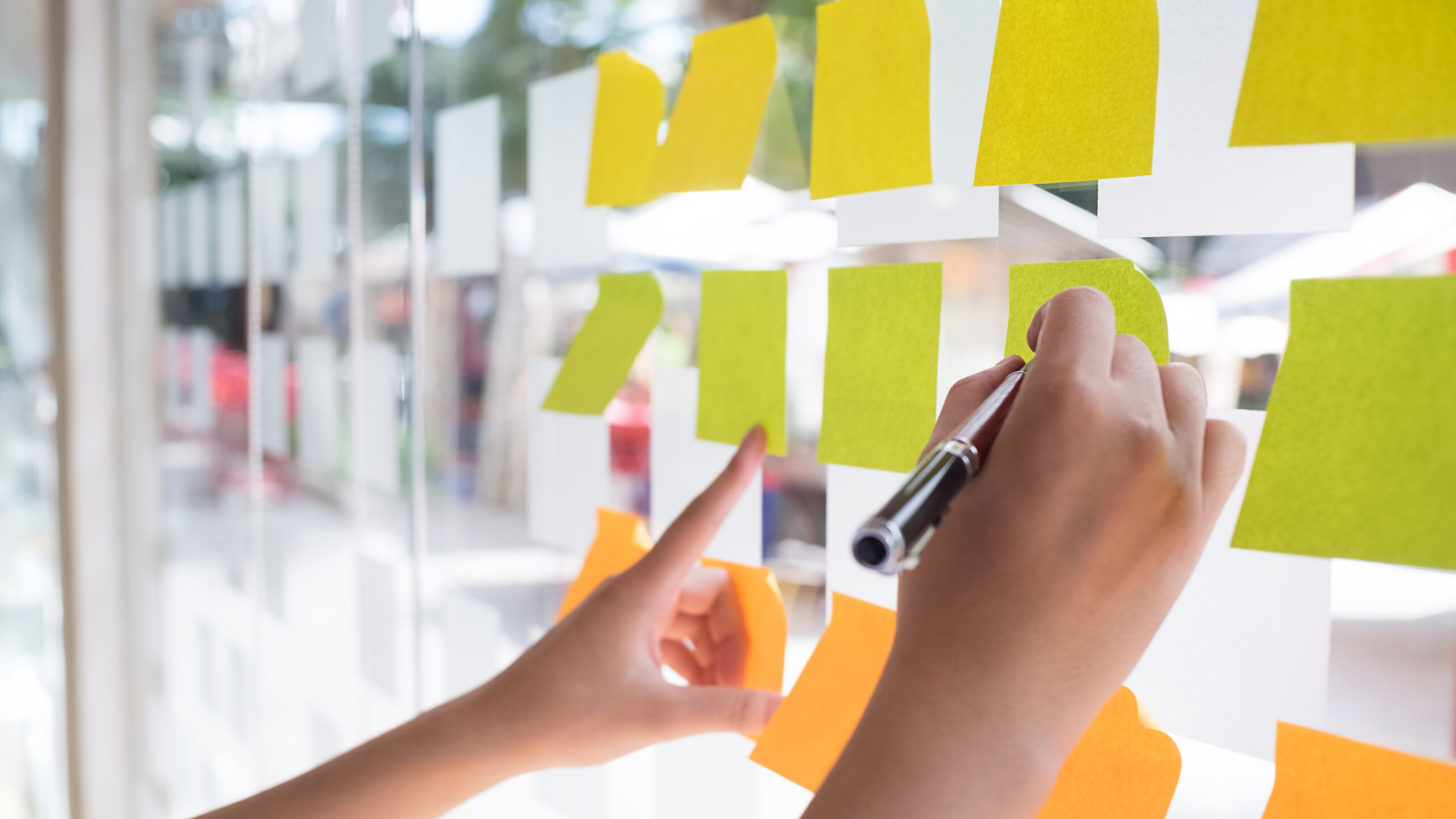 Multicoloured post-it notes are arranged on a glass window. Two hands are visible, one holding a pen as though to write on one of the notes.